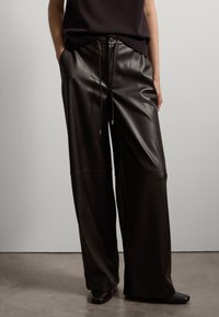 Pantalons à jambes larges en cuir brun foncé, dotés d'une taille à cordon, d'une texture lisse et de détails de couture minimaux, associés à des chaussures foncées.