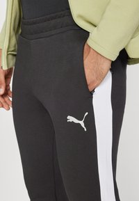 Pantaloni de training negri cu dungi albe pe lateral, având un material texturat și un mic logo Puma alb pe coapsa stângă.