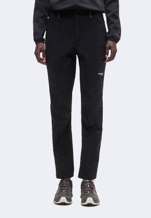 Personne portant un pantalon de plein air noir et des chaussures de randonnée grises, debout devant un fond clair uni.