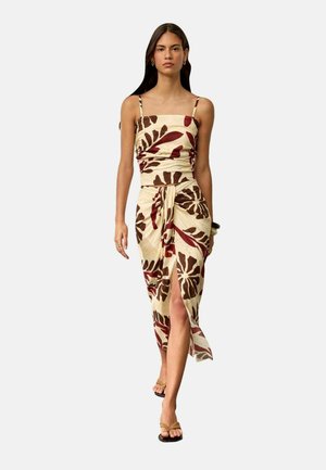 Mujer caminando con un vestido beige con patrones de hojas marrones y rojas, con tirantes finos y una abertura frontal, llevando sandalias color cuero.