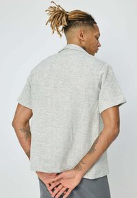 Chemise à manches courtes en tissu léger et texturé, de couleur gris clair, avec un motif subtil ; elle présente un col plat et un ourlet droit.