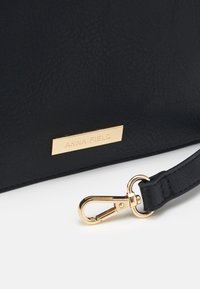 Sac à main en cuir synthétique noir avec une surface texturée, doté d'une étiquette en métal doré portant l'inscription "ANNA FIELD" et d'une fermeture à fermoir en métal doré.