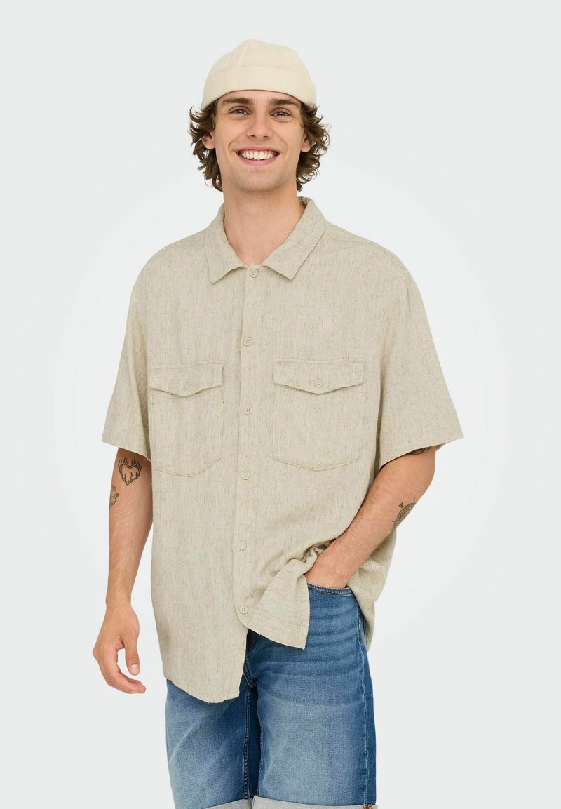 Lyse beige kortærmet skjorte med knaplukning og to brystlommer, lavet af tekstureret stof. Model iført blå denimshorts og en hvid kasket.