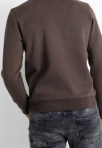 Hnědá mikina s hladkou texturou, kulatým výstřihem a žebrovaným lemem. Doplněná šedými džíny s více kapsami a vybledlým vzhledem.