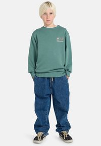 Sudadera de algodón verde con puños con ribete, que presenta el logo de Billabong; combinada con unos pantalones vaqueros azules holgados y zapatos negros con cordones beige.