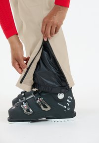 Beige Skihosen verfügen über einen schwarzen Reißverschluss an den Säumen und zeigen eine glatte Textur. Darunter sind schwarze Skischuhe mit silbernen Beschlägen abgebildet.