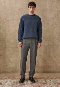 Navy knitted sweater with ribbed cuffs, paired with light gray pleated trousers and black leather shoes on a patterned rug. Neutral textured background.