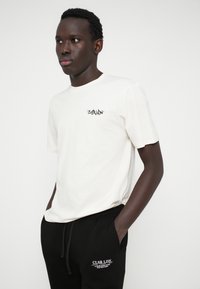 Κρεμ μπλούζα φτιαγμένη από βαμβάκι με στρογγυλή λαιμόκοψη και κοντά μανίκια, που διαθέτει ένα μικρό μαύρο λογότυπο στην αριστερή τσέπη του στήθους. Μαύρα αθλητικά παντελόνια με άσπρο λογότυπο.