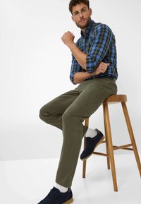 Chemise à carreaux dans des tons de bleu et de vert, pantalon vert olive et baskets navy, assis sur un tabouret en bois, avec un fond neutre et lisse.