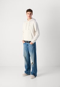 Cremefarbener Hoodie mit Kordelzügen und Fronttasche, kombiniert mit hellblauen, abgewetzten weit geschnittenen Jeans und beigen Sneakers, vor einem neutralen Hintergrund.