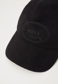 Black fleece cap featuring an oval patch with "BOSS" and "EST. 1991" embossed. The design includes textured fabric and stitching details.