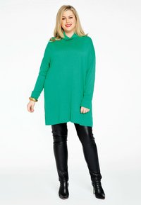 Groene oversized tuniektrui met een col, lange mouwen en een losse pasvorm, gecombineerd met zwarte leren legging en puntige enkellaarzen.