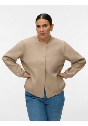 Sötét hajú nő bézs színű, gombos dzsekiben és kék farmernadrágban, kezeit csípőjén tartva egy sima fehér háttér előtt áll.