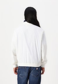 White bomber jacket with elastic cuffs and hem, made of smooth fabric; worn with blue jeans featuring a small logo on the back pocket.