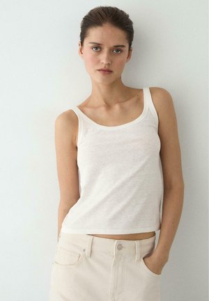 Frau mit heller Haut und dunklem Haar, die ein weißes ärmelloses Oberteil und hellfarbene Hosen trägt, mit einer Hand in der Tasche, vor einer weißen Wand stehend.