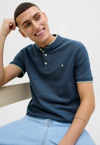 Marineblaues Poloshirt mit Kragen, drei Knöpfen und hellgrünen Akzenten. Trägt dazu hellblaue Jeans und sitzt auf einer Holzbank.
