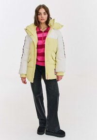 Dúková bunda v žltej a bielej farbe, so zipsom, veľkými vreckami a kapucňou. Nosená cez ružový pruhovaný vrch s čiernymi nohavicami.