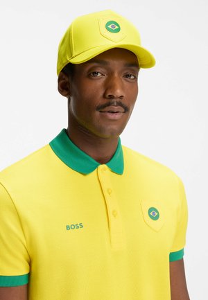Homme portant une casquette jaune et un polo avec un bord vert, orné de patchs du drapeau brésilien et du logo "BOSS" sur le polo.