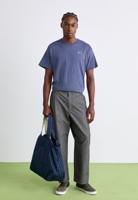 Ung mand med fletninger iført en blå Nike T-shirt, grå bukser og grønne sneakers, som holder en blå mønstret tote taske.