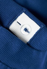 Tissu bleu marine avec une texture en côtes, comportant une étiquette blanche et un détail de bouton transparent attaché à la couture du col.