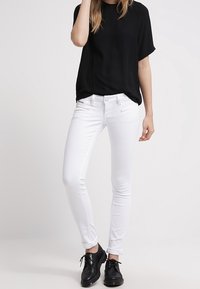 White slim-fit jeans with a smooth texture, five pockets, and belt loops, paired with a loose black short-sleeve top and black shoes.