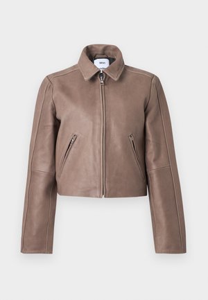 Žena s okuliarmi na slnko, taupe kožená bunda, čierny rebrovaný top, široké modré džínsy, čierne špicaté podpätky, stojaca pred jednoliatym pozadím.