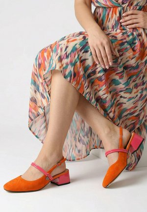 Vrouw zit met gekruiste benen, draagt een kleurrijke jurk met patroon en oranje schoenen met blokhak en roze bandjes.