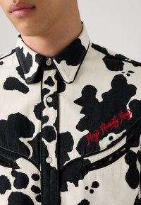 Hvit denimskjorte med svart kuemønster, spiss krage, to brystlommer, og "Hey Howdy Hey" brodert i rødt på venstre side.