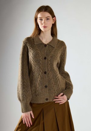 Jonge vrouw draagt een bruine kabelgebreide cardigan met knopen en bijpassende bruine geplooide rok, staand tegen een effen achtergrond.