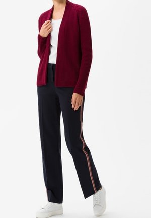 Cardigan rodato color bordeaux con design aperto, indossato sopra un top bianco. Abbinato a pantaloni blu navy con strisce laterali rosse e bianche e sneakers bianche.