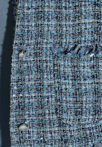 Blauw tweed stof met een ruitpatroon, met een gestructureerd oppervlak en zilveren knoopaccenten. Toont een zakdetail en rafelige randen.