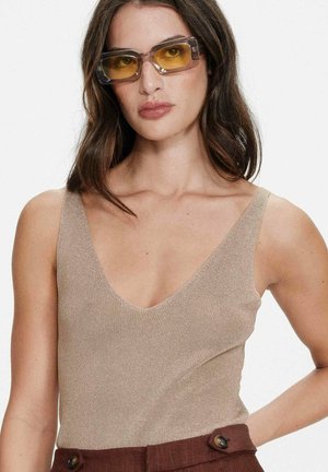 Femme portant un haut beige sans manches à col en V, un pantalon marron taille haute avec des boutons, et des lunettes de soleil rectangulaires aux verres jaunâtres, posant devant un fond blanc.