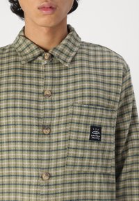 Short-sleeve plaid shirt in green and beige tones, featuring a button-up front, rounded collar, and a chest pocket with a black logo patch.