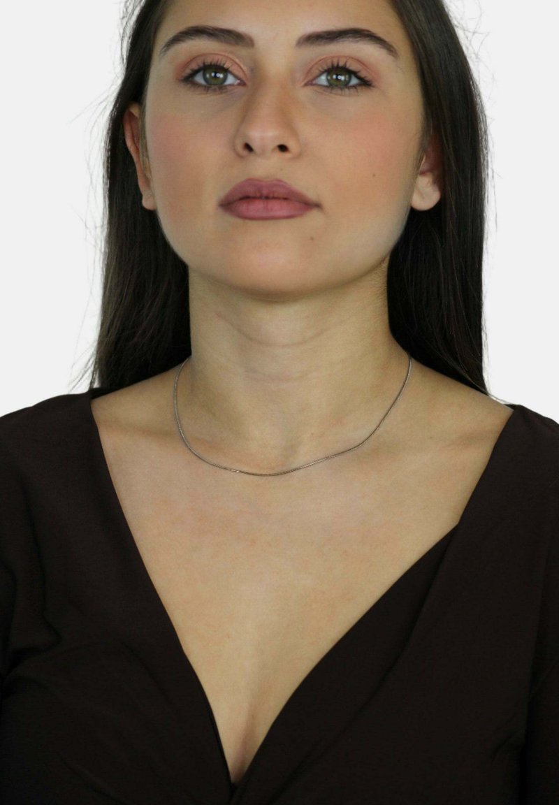 Srebrny wężowy naszyjnik spoczywa na nagim obojczyku, charakteryzując się gładką, błyszczącą powierzchnią oraz elastycznym, cienkim wzornictwem na tle ciemnej bluzki.