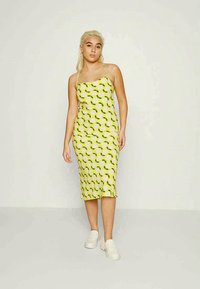 Vestido midi sem mangas amarelo com um padrão de folhas, apresentando uma silhueta ajustada e uma textura suave. Combinado com ténis brancos.