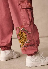 Rosa cargobyxor med broderad tiger och lövdetalj; elastiska muddar; logotyp "Superdry" i kursiv stil; kombinerat med vita sneakers.