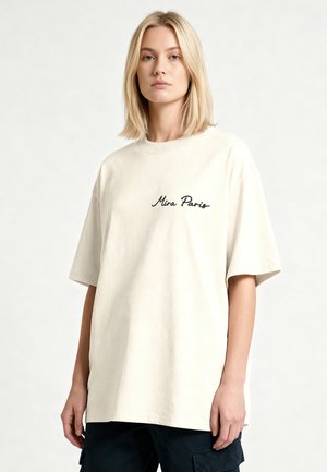 Femme blonde portant un T-shirt oversize blanc cassé avec le texte "Mira Paris" et un pantalon cargo noir sur un fond clair uni.