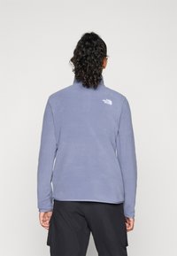 Cilvēks ar cirtainiem matiem, sasietiem atpakaļ, ģērbts gaiši zilā flīsa jaka ar The North Face logo, stāv pretim vienkāršai fona.
