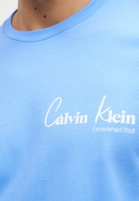 Κοντινό πλάνο ενός ατόμου που φοράει ανοιχτό μπλε πουκάμισο με λευκό κείμενο "Calvin Klein Established 1968" στο στήθος.