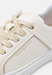 Białe skórzane sneakersy z teksturowanymi złotymi sznurówkami i subtelnymi przeszyciami. Posiadają logo "TOMMY HILFIGER" z boku oraz białą podeszwę.