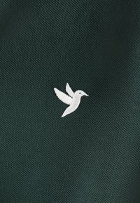 Tissu vert foncé avec un petit oiseau brodé blanc aux ailes déployées, sur un fond texturé.