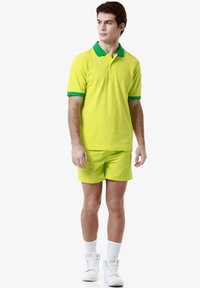 Polo shirt giallo neon con colletto e polsini verdi, abbinato a pantaloni corti coordinati. Indossato con sneakers bianche e calze. Tessuto dalla trama liscia.