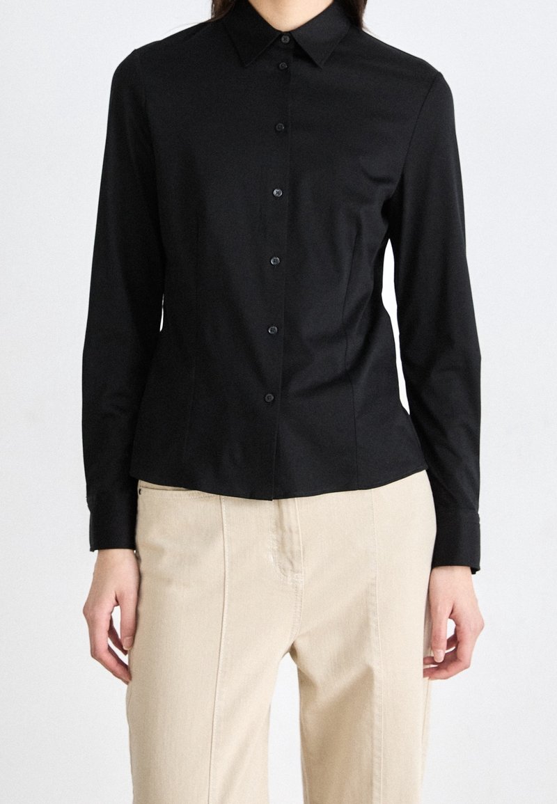 Femme portant une chemise noire cintrée à manches longues boutonnée, associée à un pantalon beige taille haute, debout devant un fond uni.