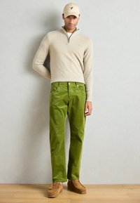 Beige zippered sweater, grønne fløjlsbukser, brune slip-on sko og en lys beige kasket med et lille grønt logo. Flade, strukturerede overflader.