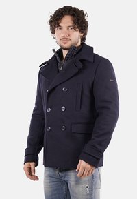 Cappotto doppiopetto navy realizzato in tessuto resistente, con grandi bottoni, colletto a punta e tasche laterali; fodera interna testurizzata visibile.
