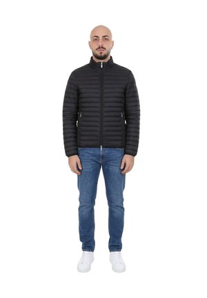 Uomo in piedi di fronte, con giacca imbottita nera, jeans blu e scarpe da ginnastica bianche su sfondo bianco.