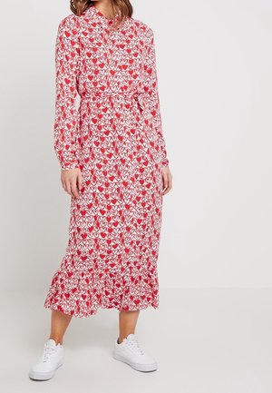 Kobieta w długiej sukience z długim rękawem sięgającej do kostek, w czerwono-biały wzór serc, z paskiem w talii i białymi trampkami.