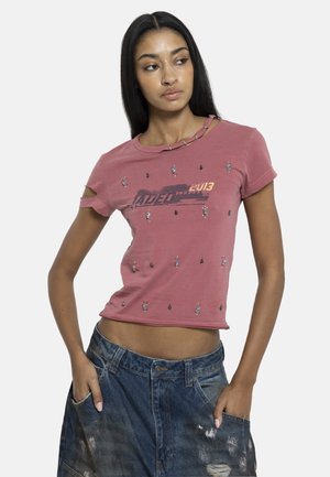 Νεαρή γυναίκα που φοράει ροζ διακοσμημένο T-shirt με κομμένους ώμους και φθαρμένο τζιν, ποζάρει με τα χέρια στις τσέπες κοιτάζοντας δεξιά.