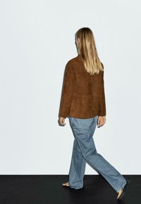 Bruine suède jas met een relaxed fit, gecombineerd met lichtblauwe rechte jeans, blootsvoets gedragen tegen een minimalistische achtergrond.