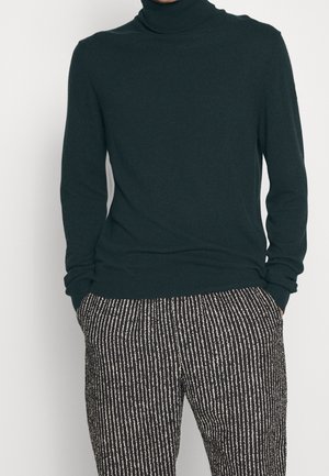 Persona che indossa un dolcevita verde scuro a maniche lunghe e pantaloni a righe verticali bianche e nere, con le mani nelle tasche.